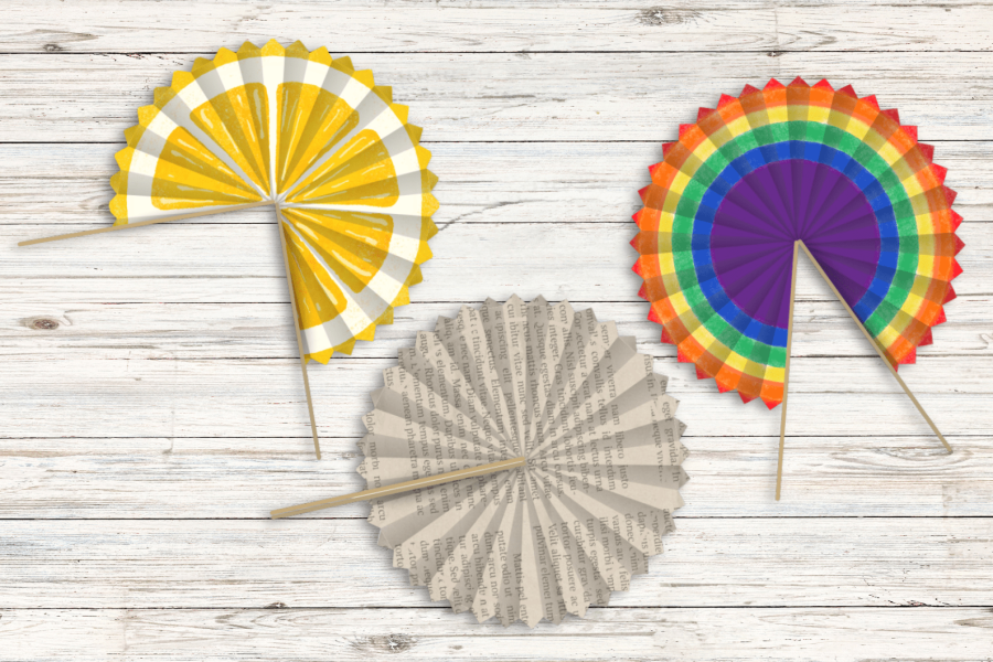 Three folded handfans on a wood table. One is made of a book page, one has a lemon design, one has a rainbow design.